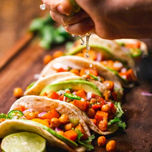 Hands pour salsa onto tacos loaded with chopped vegetables and herbs, with lime wedges beside them on a rustic table.