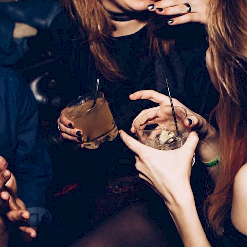 Two people at a night out, holding cups with straws and sipping dark drinks, with bold nail art and tattoos visible.