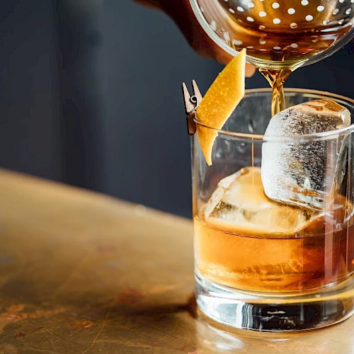 Amber cocktail with ice is being poured into a rocks glass, garnished on a skewer, on a bar counter.
