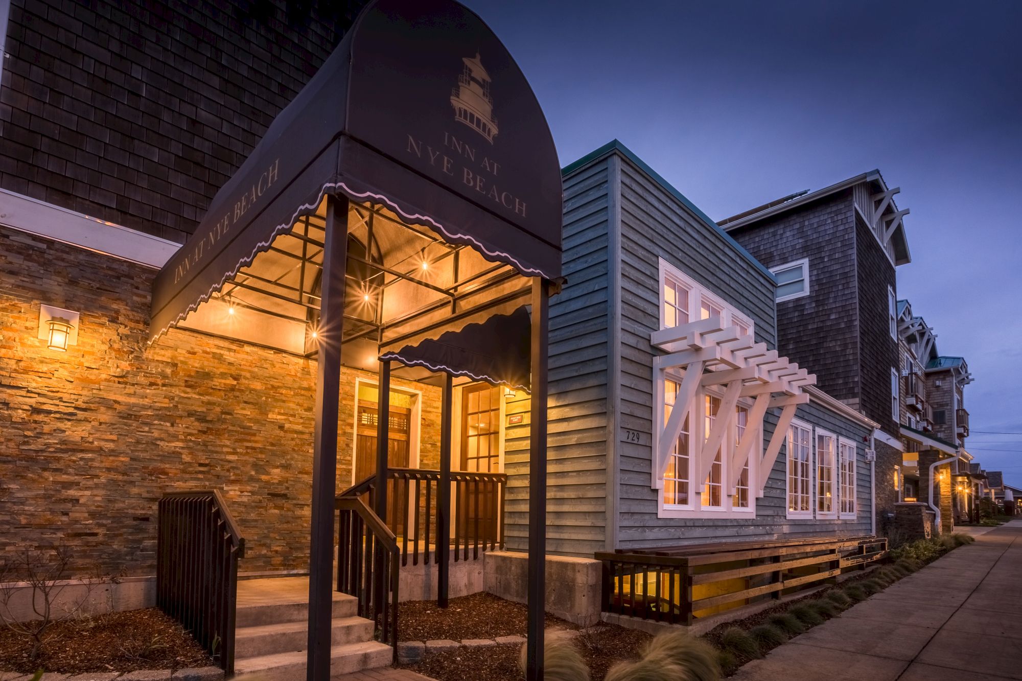 A cozy storefront with warm lights under a curved awning, stone and wood exterior, white decorative trim, and a quiet evening street.