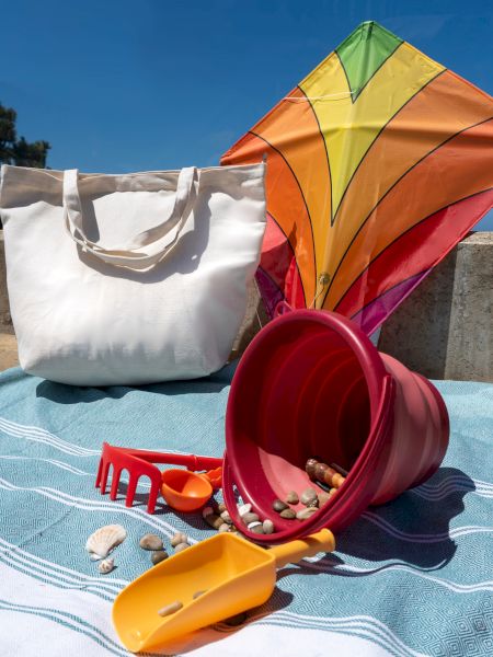 A colorful diamond kite beside a white beach tote, with a red bucket, sand tools, shells, and pebbles on a blue blanket.