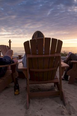 A group of people sits in wooden beach chairs around a small fire at sunset on the sand, enjoying drinks and conversation by the ocean.