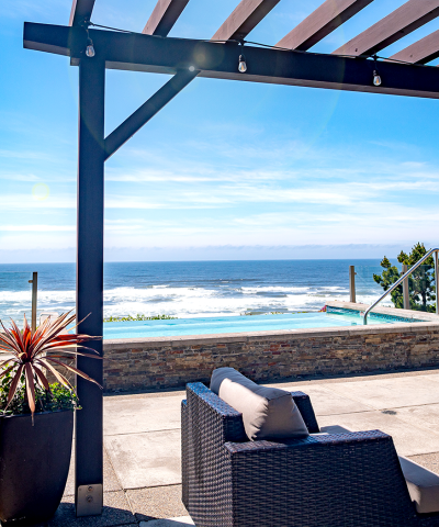 A sunny beachfront patio with a pergola, wicker furniture, a stone planter, and an ocean view with waves beyond a low wall.