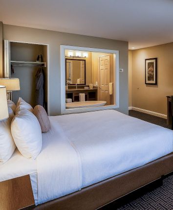 A cozy hotel room with a large bed, nightstands, a desk, a wall-mounted TV, a separate sitting area, and a fireplace, all in warm, neutral tones.