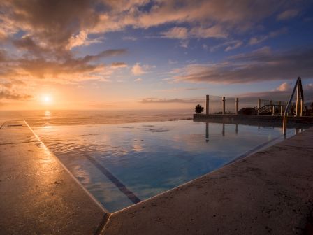 A tranquil outdoor pool reflecting a sunset over calm water, with a pier, steps, and a dramatic orange-pink sky.