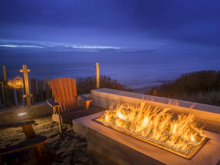 A nighttime patio scene with a glowing rectangular fire feature, Adirondack chair by a railing, and blue dusk sky over dunes in the background.
