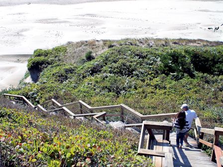 A wooden stairway winds down a grassy dune to a sandy beach; two people stand at a railing enjoying the view.