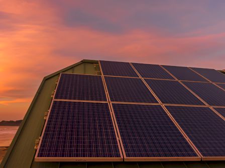 Solar panels on a slanted roof at sunset, golden sky, calm beach in the background, warm hues reflecting off the panels.