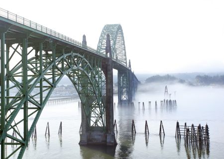 A green steel truss bridge spans a foggy river, with piers in calm water and old bridge sections in the distance.