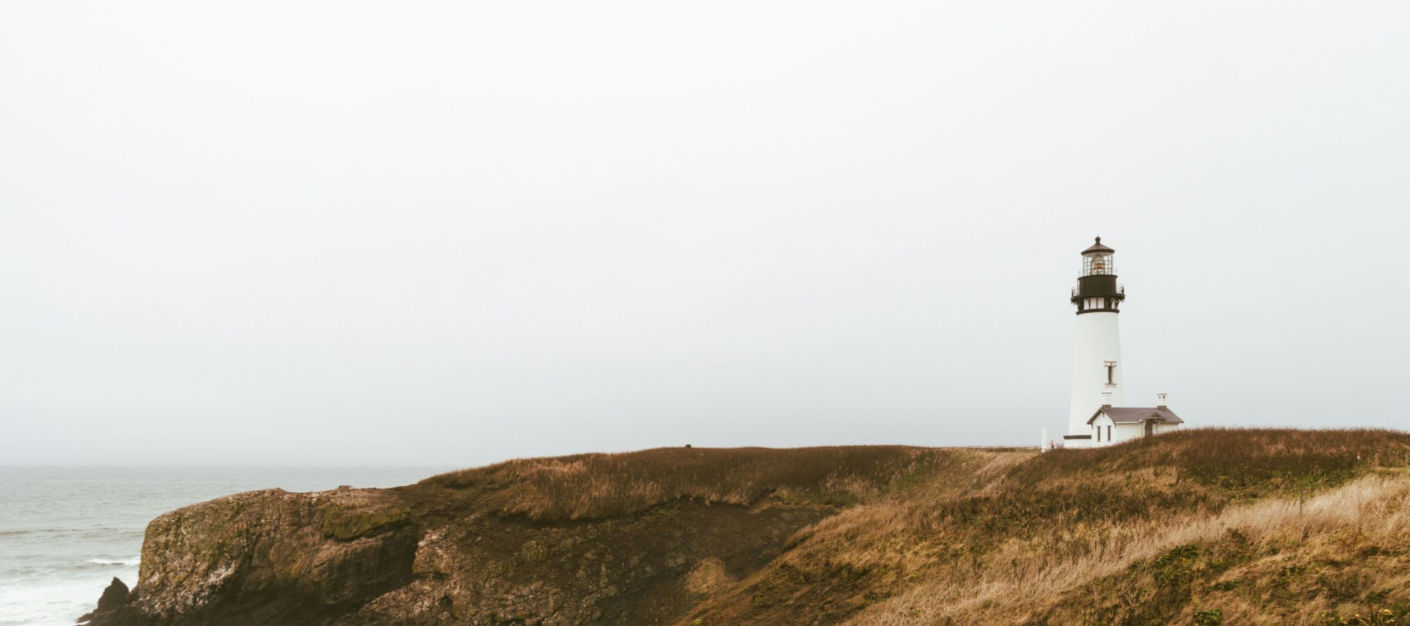 A tall white lighthouse stands on a grassy cliff by the ocean under a pale sky.