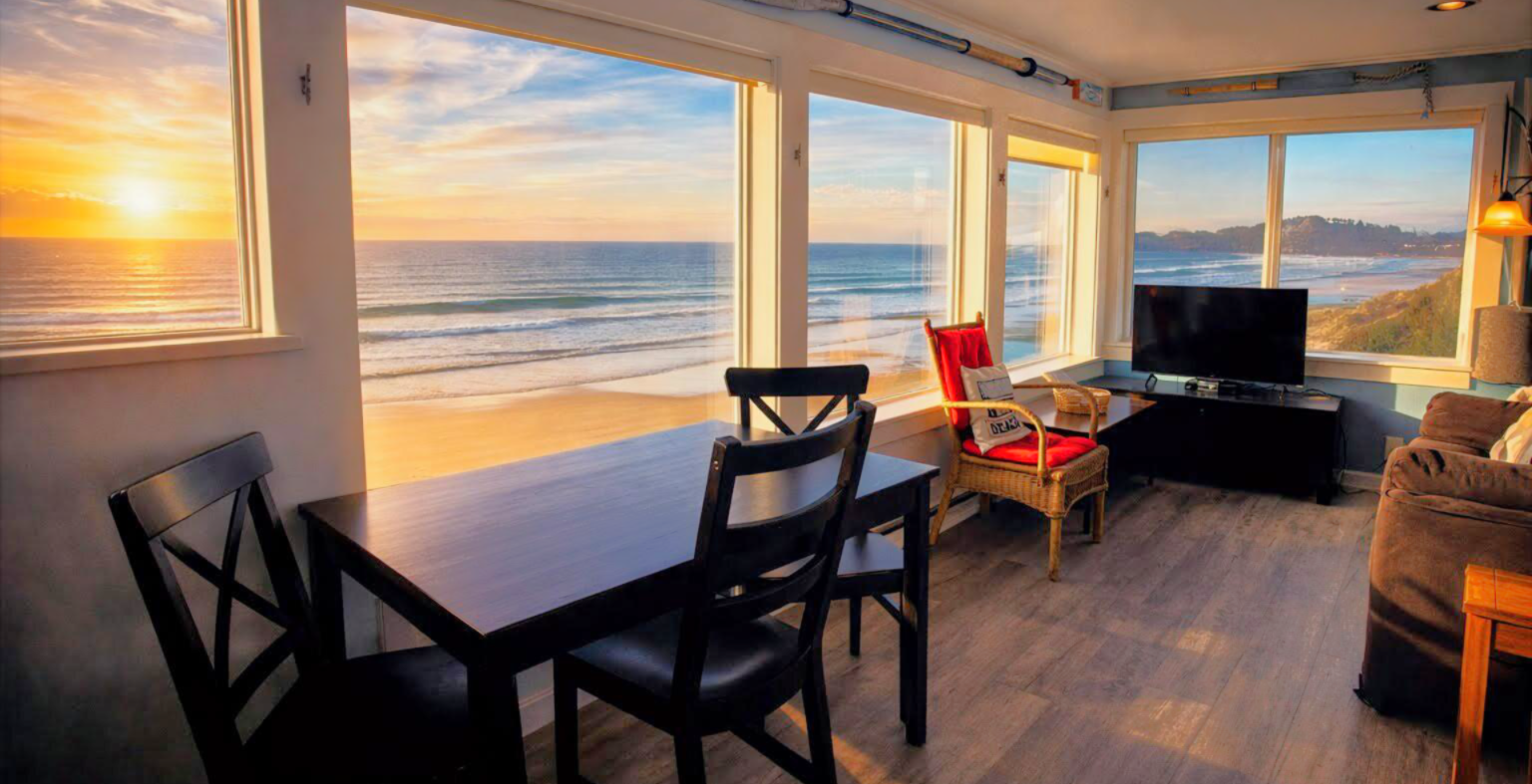 A cozy seaside living room with dining table, chairs, TV, and large ocean-view windows at sunset.