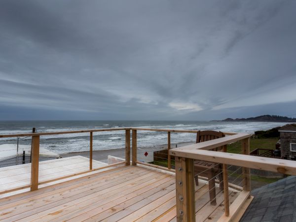 A wooden deck overlooks a gray ocean with waves and distant coastline under cloudy skies.