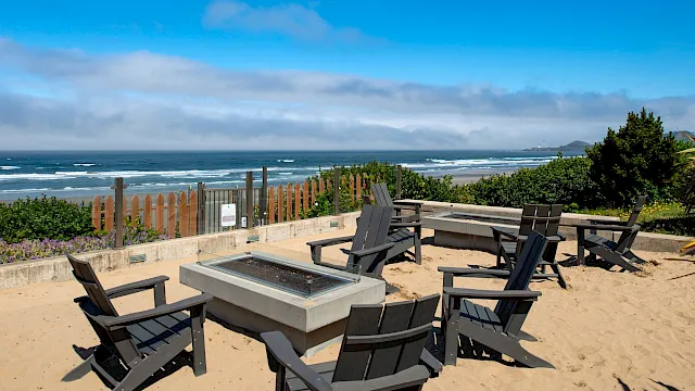 A beachside fire pit area with eight black chairs arranged around a square fire pit, sandy ground, dunes, and the ocean in the background; sunny skies.