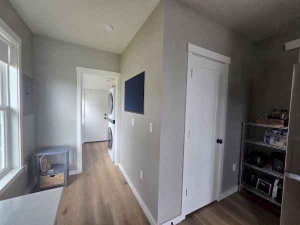 A modern laundry/broom area with white paneled doors, light wood floors, a window, and open shelving with kitchen items.