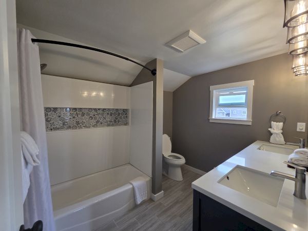 Bright modern bathroom with a bathtub, tiled shower accent wall, toilet, vanity sink, window, and ceiling vent.
