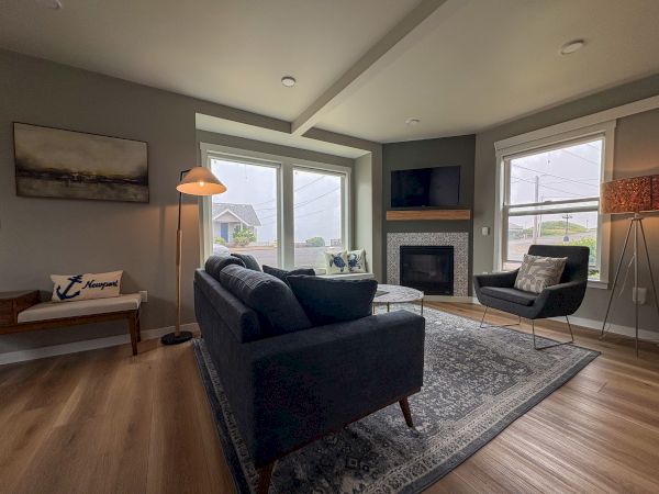 A cozy living room with dark couches, a patterned rug, fireplace, two windows, wall art, and floor lamp with glass shades.