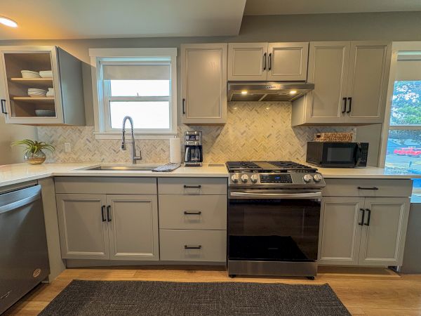 Bright modern kitchen with white cabinets, tiled backsplash, sink under window, and stainless stove, hood, microwave, and coffee maker.