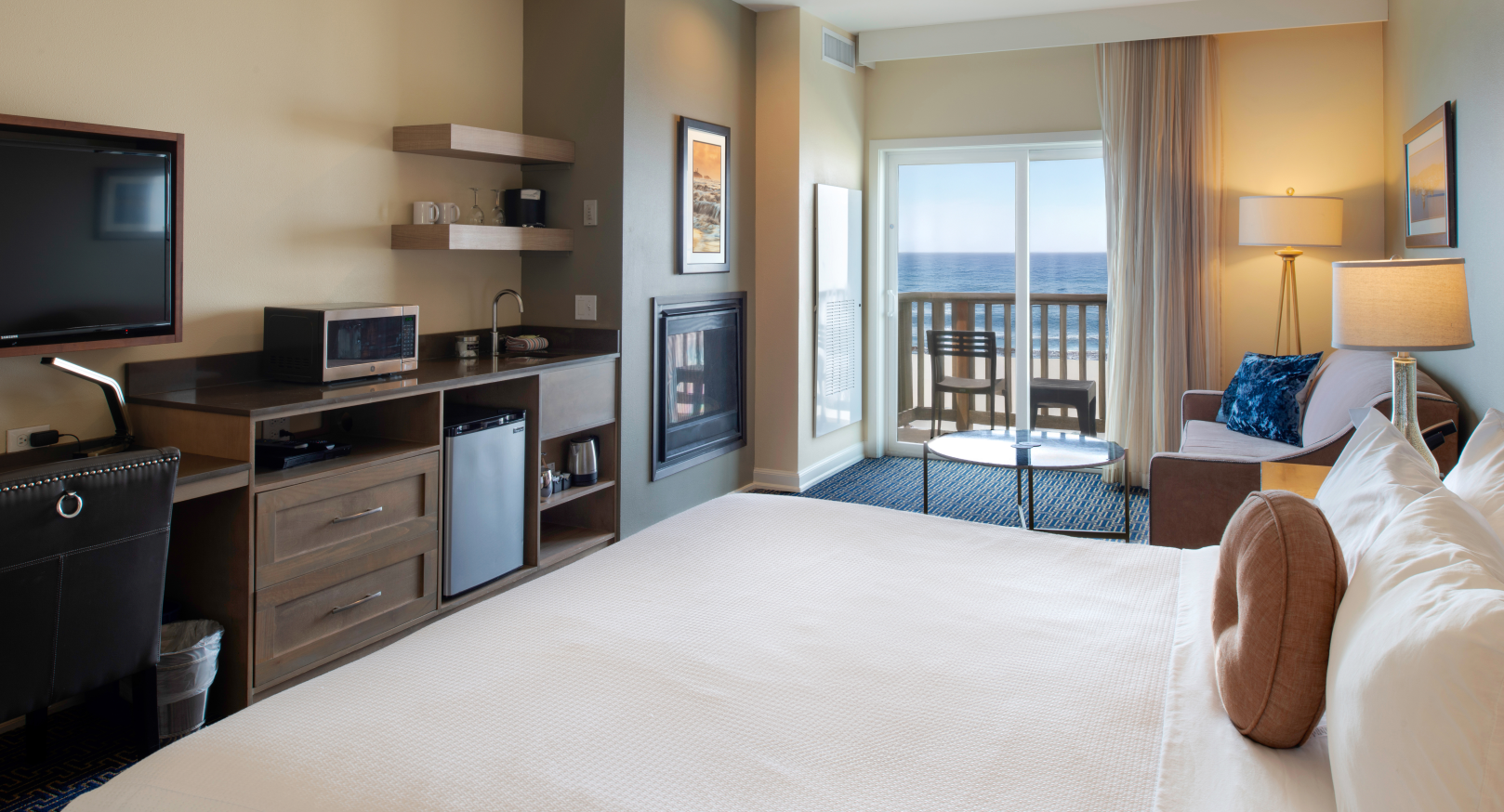 A modern hotel room with a large bed, small kitchenette, and ocean view through sliding doors, lit by warm lamps.
