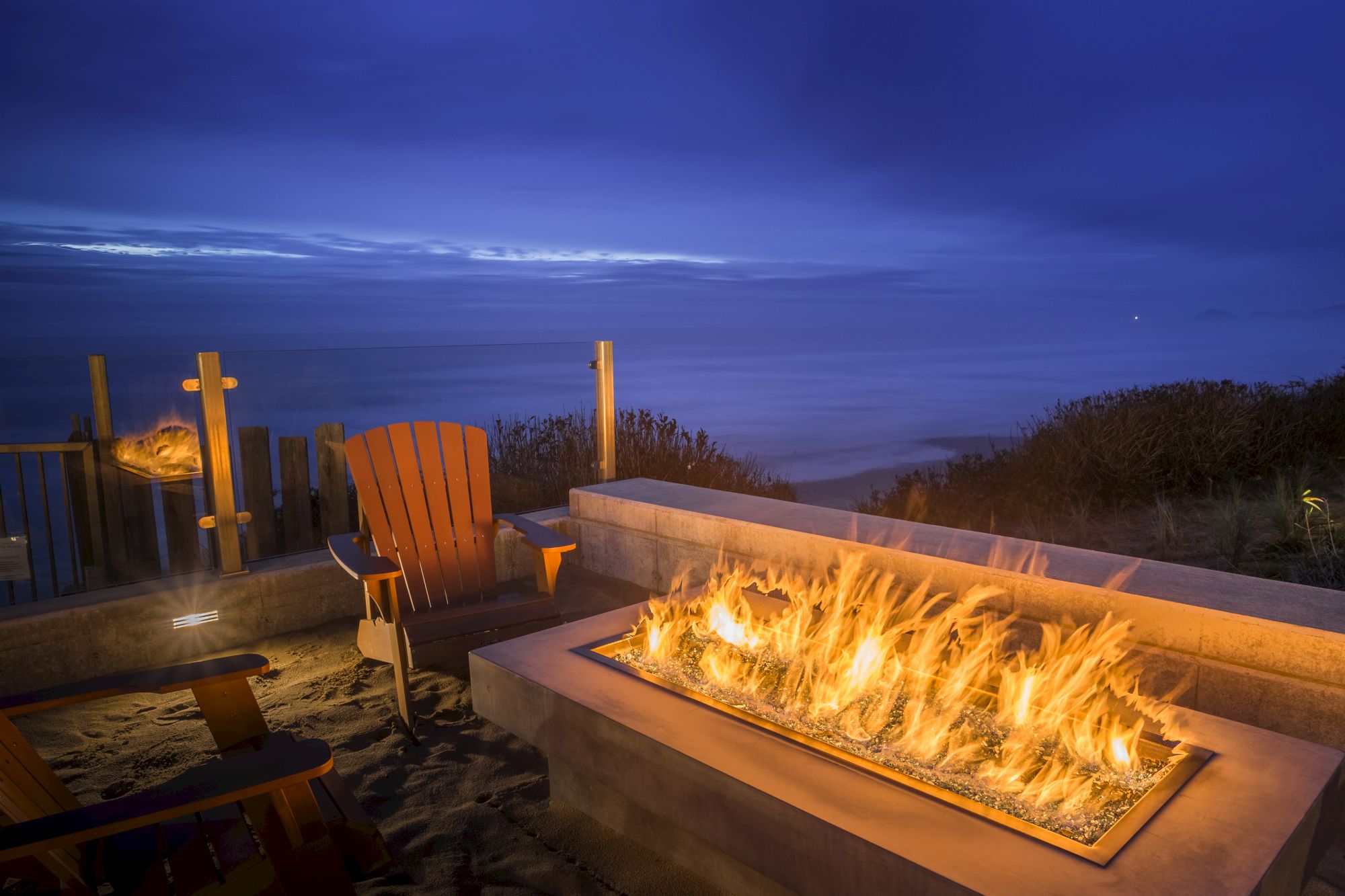 A beach patio at dusk with a large outdoor fire pit blazing and Adirondack chairs facing the sea.