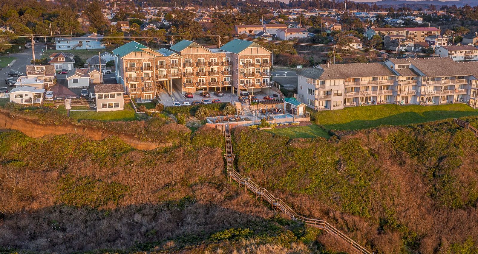 Aerial view of a residential apartment complex with parking lots, fenced patios, grassy hills, and a distant town at dusk.