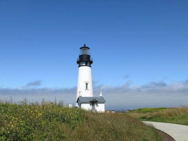 A white lighthouse with a dark lantern room rises above grassy coastal hills under a blue sky with clouds.